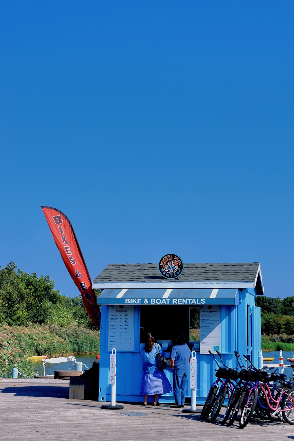 Blue bike and boat rental kiosk on a park boardwalk under a clear sky
