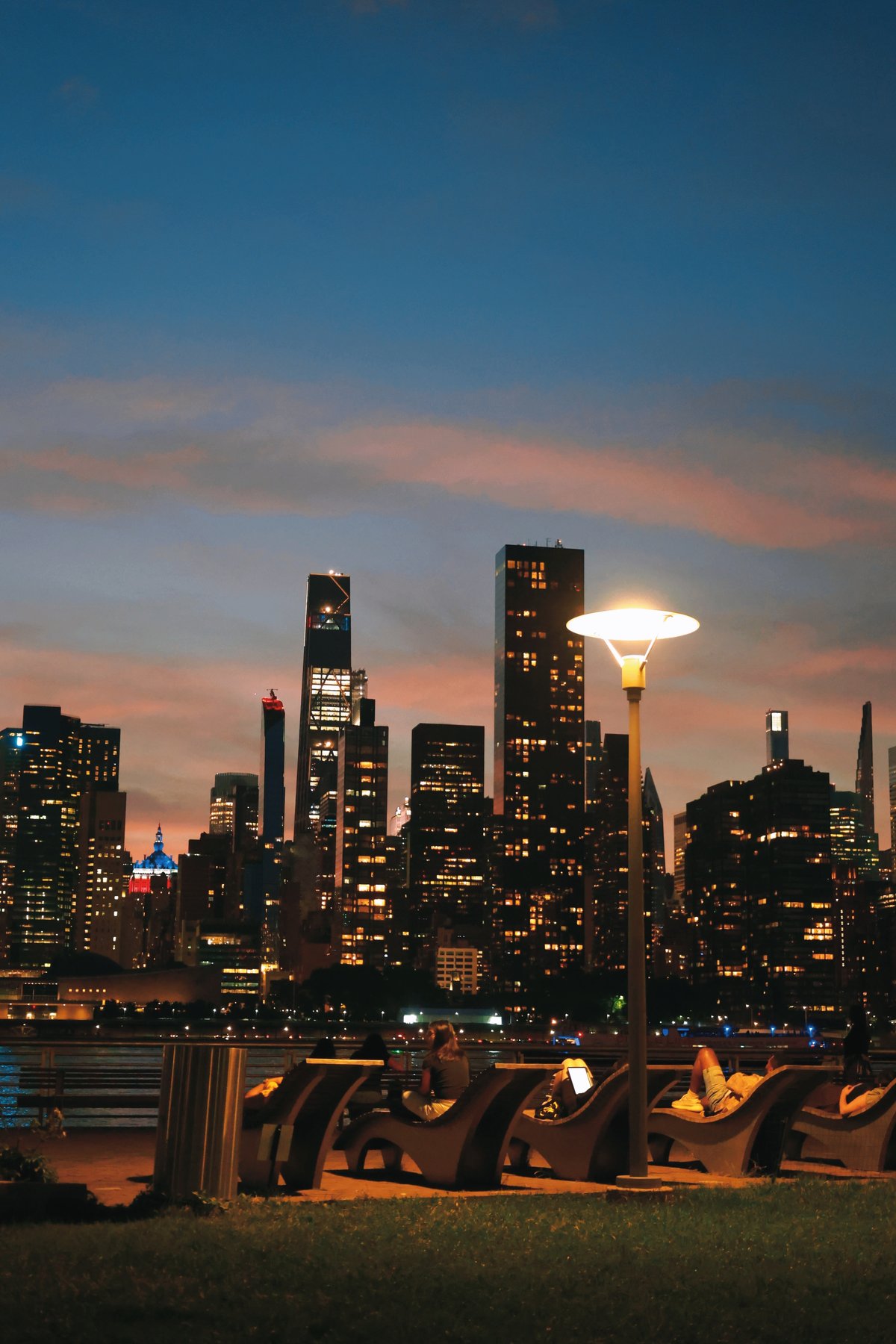 New York City skyline at dusk with people relaxing on riverside benches